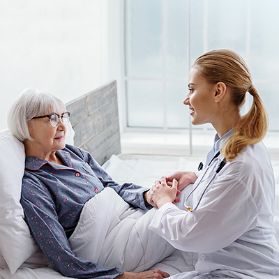 female nurse holding hands and chatting with elderly female resident sitting in bed